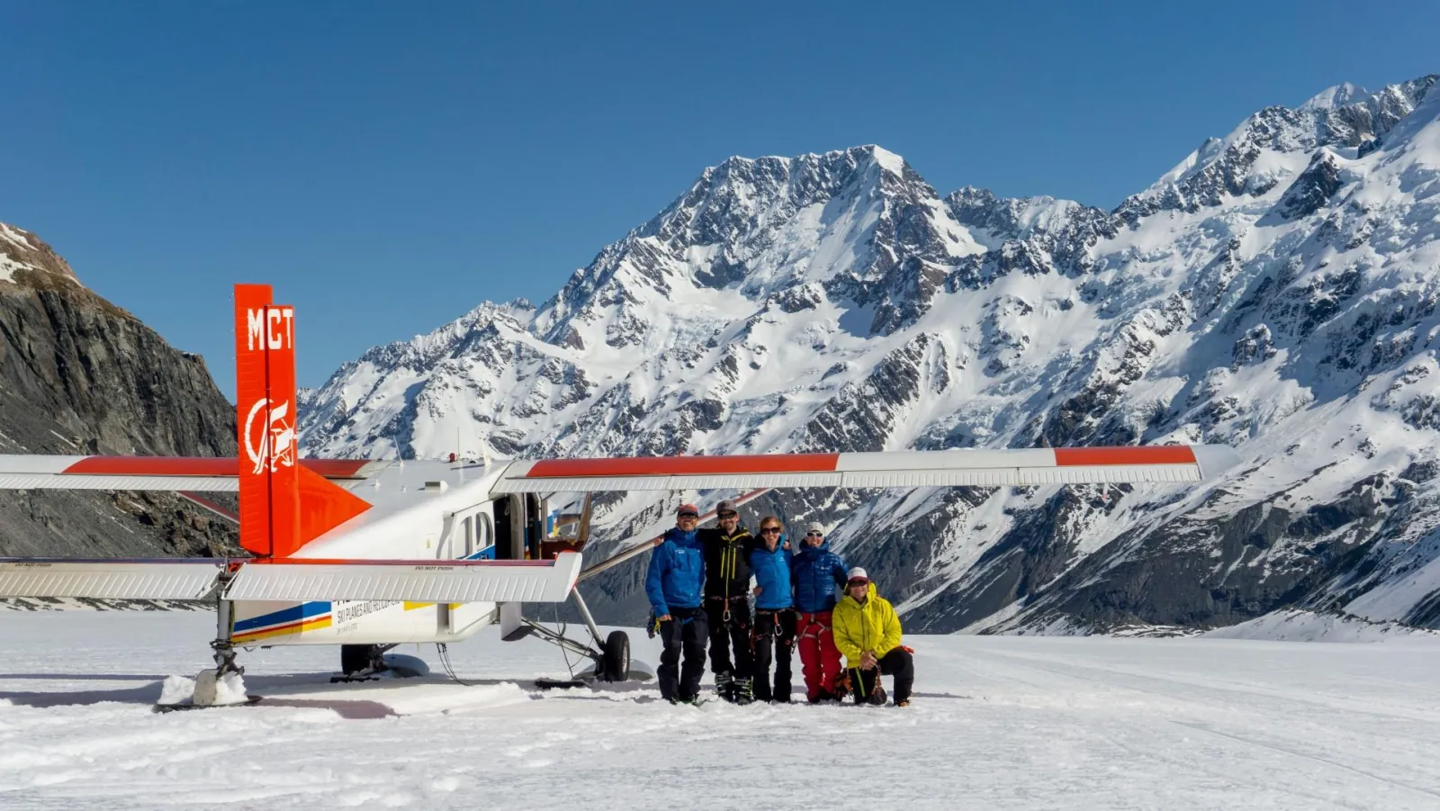 Group posing beside a red-tailed ski plane on the snow with high peaks behind