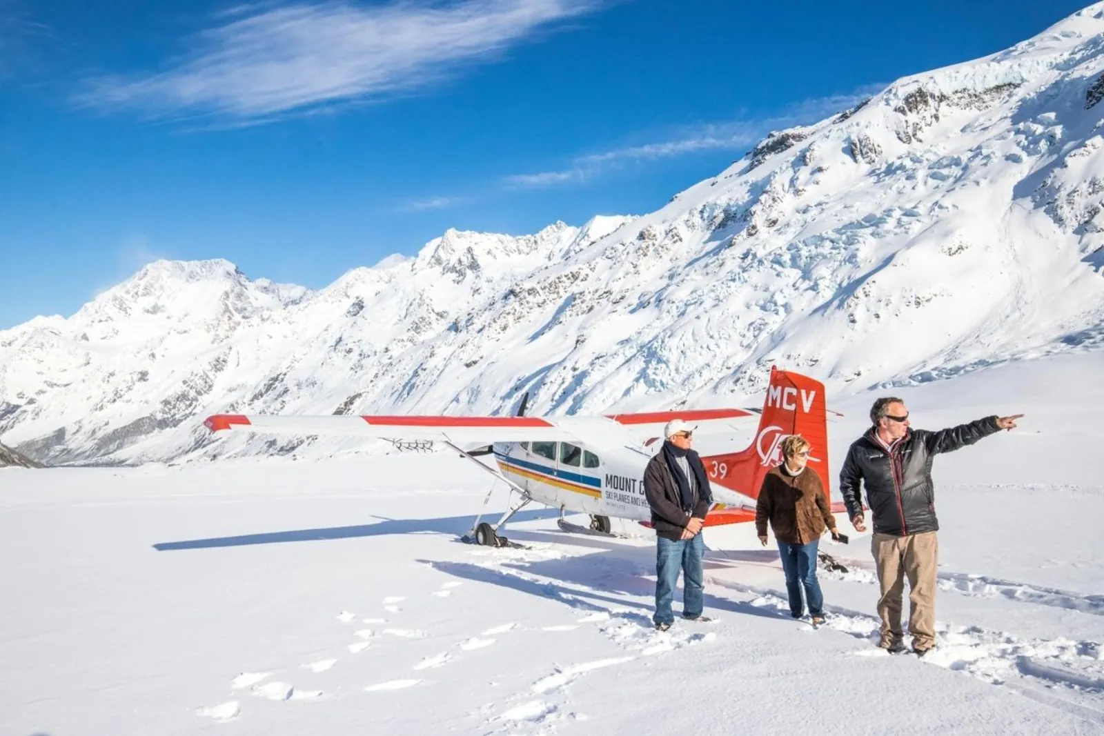 Pilot pointing out peaks to two passengers beside a ski plane on the glacier