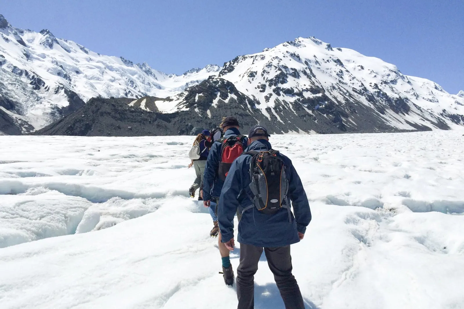 Hikers walking in single file across the glacier toward snow-capped mountains