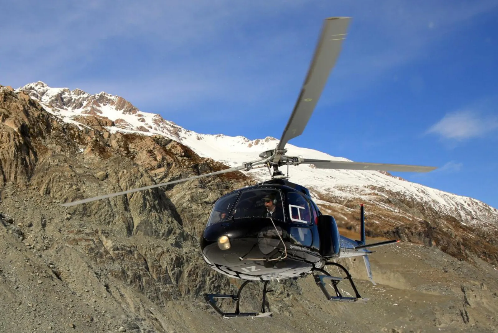 Helicopter lifting off from a rocky mountainside below snowy ridges