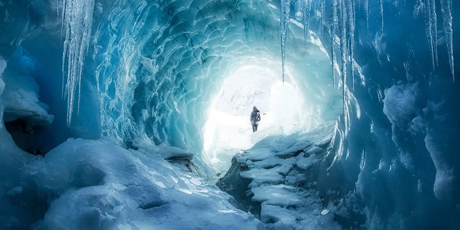 Hiker at the end of a blue ice cave with icicles hanging from the roof
