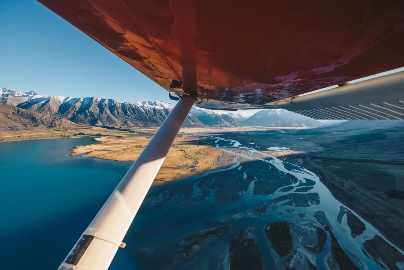 Wing strut of a ski plane framing a turquoise lake and braided river below