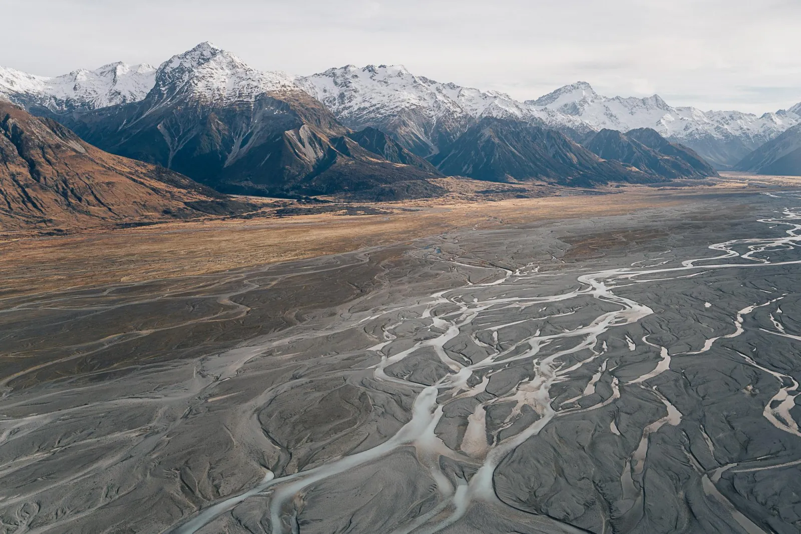 Braided river channels spreading across a valley floor below snowy mountains