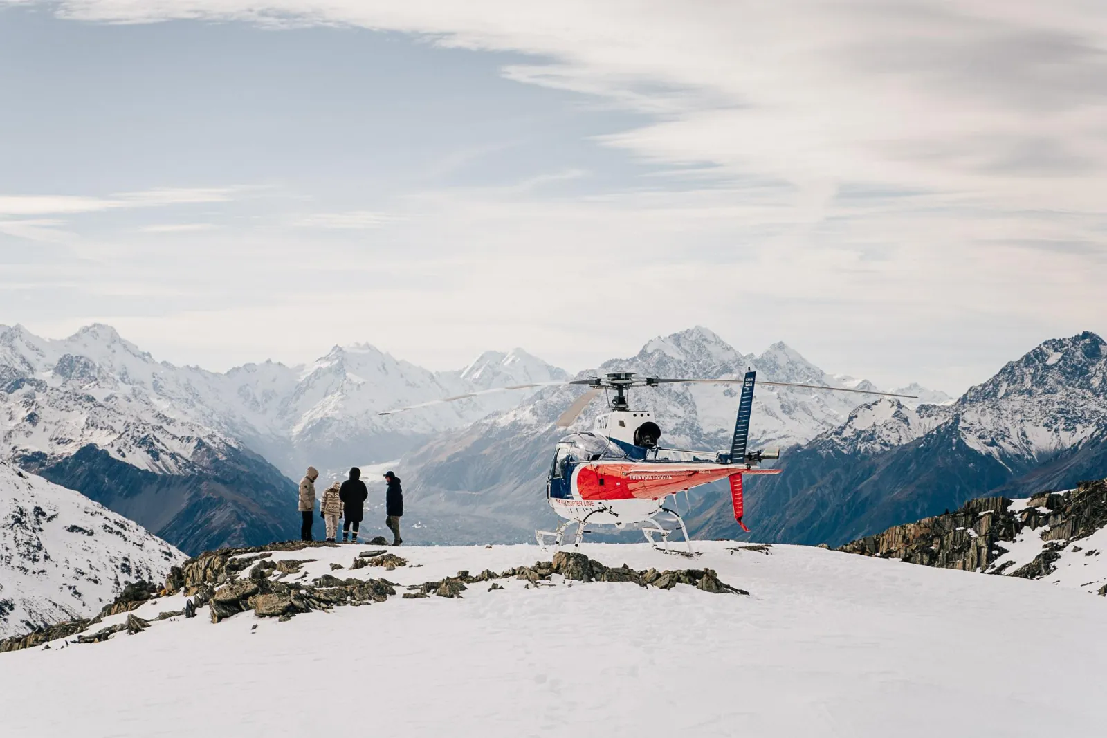 Passengers on a snowy ridge beside the helicopter, looking toward the Southern Alps