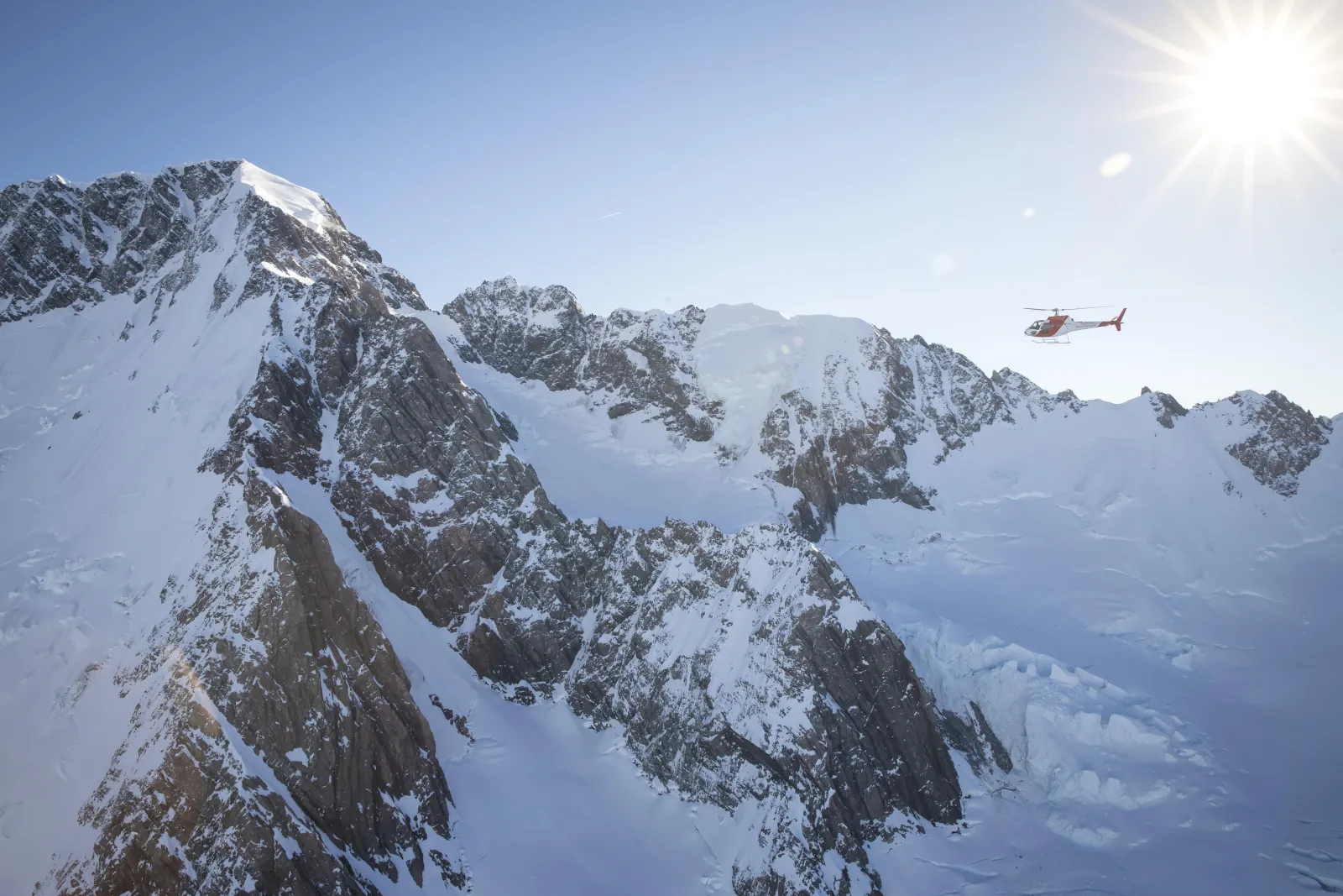 Helicopter small against the sun above a snowy mountain massif